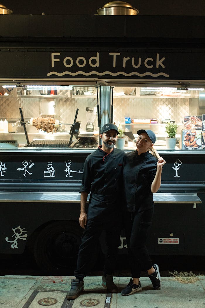 A man and woman stand in front of a food truck, offering street food at night.